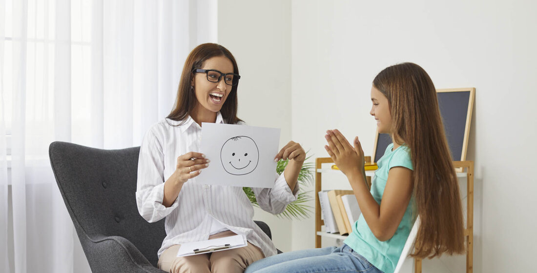 Professional school psychologist talks about positive emotions during meeting with preteen girl. Friendly female counselor shows teenage girl sheet of paper with smiley emoticon drawn on it.