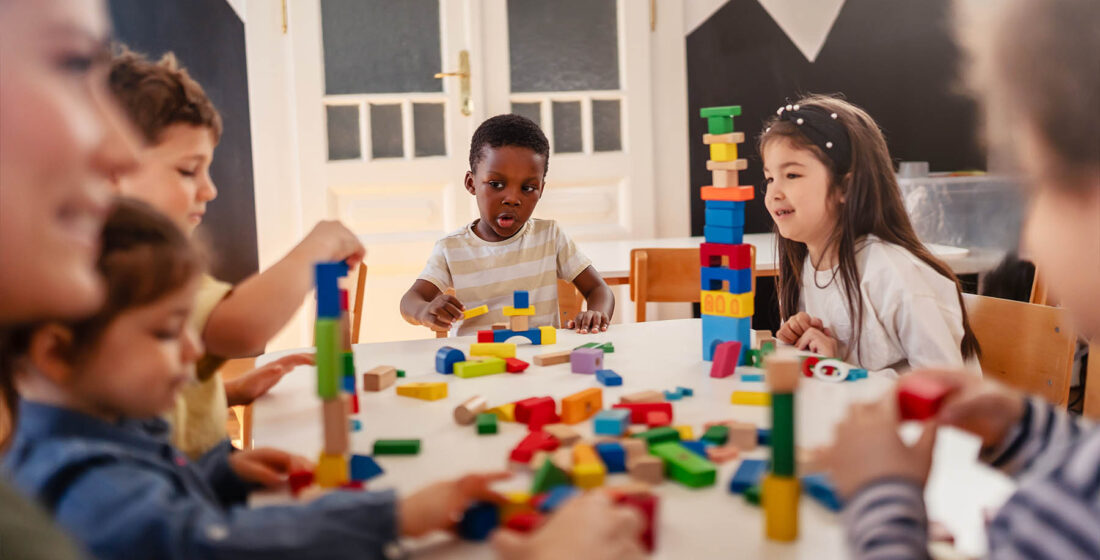 Diverse preschool children play with colorful building blocks at a classroom table, guided by a smiling teacher in a creative, inclusive early learning environment.