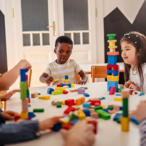 Diverse preschool children play with colorful building blocks at a classroom table, guided by a smiling teacher in a creative, inclusive early learning environment.