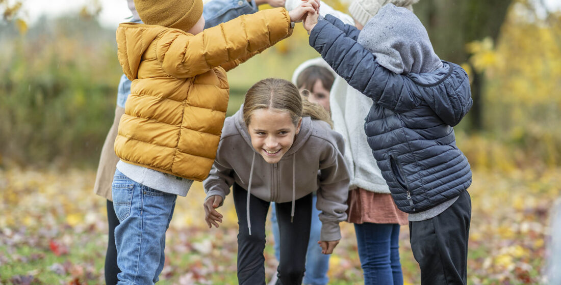 A small group of school-aged children play a game of London Bridges as they hold hands and run under the tunnel of arms. They are each dressed in warm fall layers and they enjoy the crisp Autumn air.