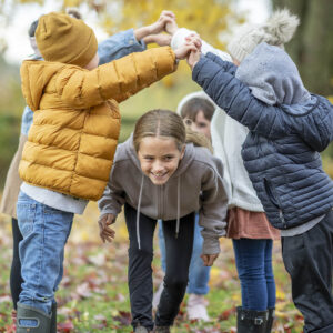 A small group of school-aged children play a game of London Bridges as they hold hands and run under the tunnel of arms. They are each dressed in warm fall layers and they enjoy the crisp Autumn air.